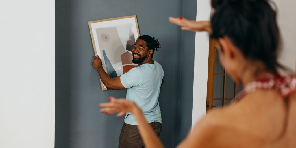 Black man holding framed picture while Caucasian woman is helping to level it against a the wall